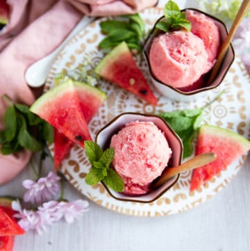 top view of two cups of watermelon sorbet in white bowls surrounded by watermelon wedges and mint leaves on a white board