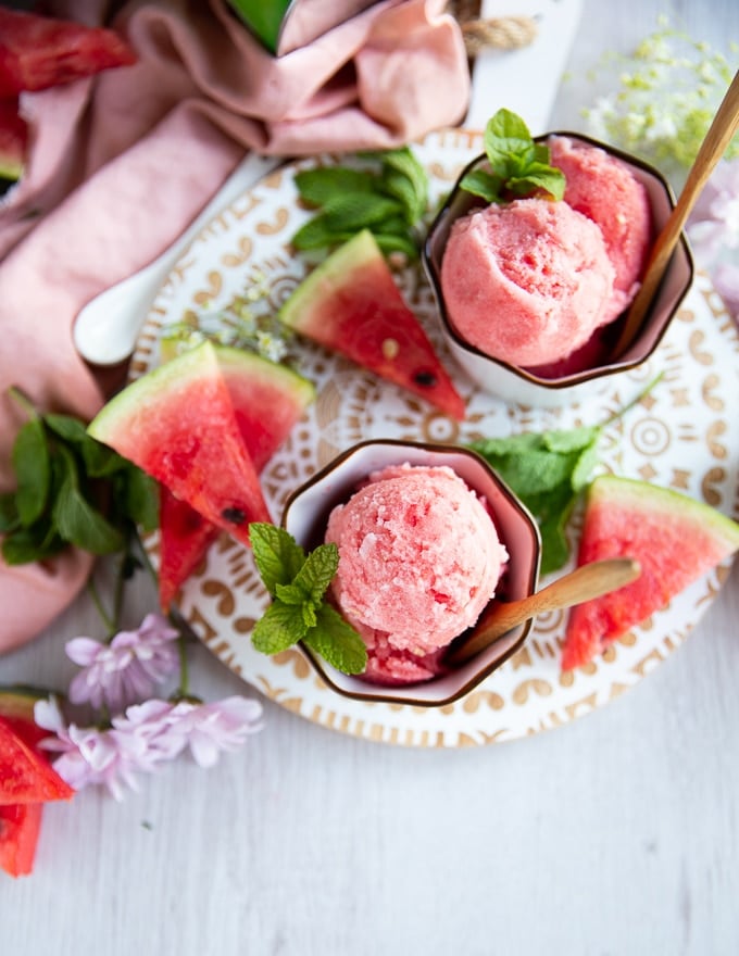 top view of two cups of watermelon sorbet in white bowls surrounded by watermelon wedges and mint leaves on a white board