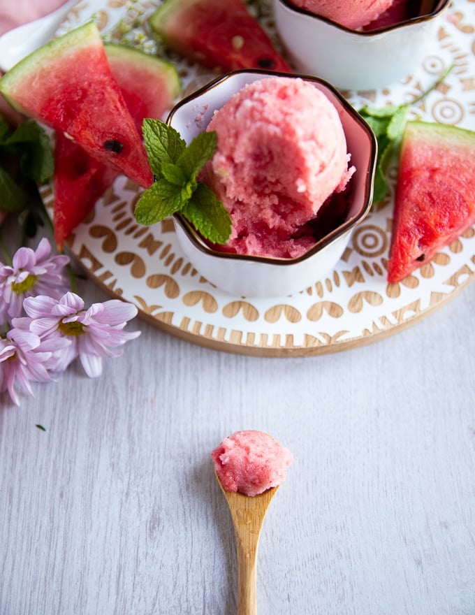 a wooden spoon with some watermelon sorbet on the board and a bowl of sorbet at the back 