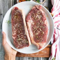 A hand holding a plate with two pieces of ny strip steak seasoned with salt, pepper, onion flakes and rosemary, ready for the grill