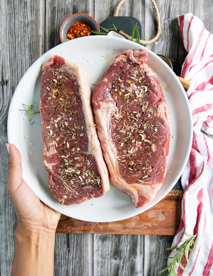 A hand holding a plate with two pieces of ny strip steak seasoned with salt, pepper, onion flakes and rosemary, ready for the grill