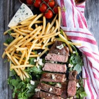 grilled NY strip steak on a board surrounded by fries, greens and a chunk of blue cheese