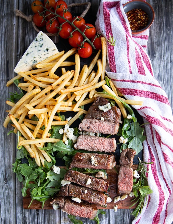 grilled NY strip steak on a board surrounded by fries, greens and a chunk of blue cheese