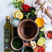 ingredients for chimichurri recipe on a board includes fresh garlic, fresh parsley, fresh oregano, salt, vinegar, lemon slices, red chillies and lots of olive oil