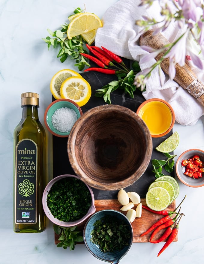 ingredients for chimichurri recipe on a board includes fresh garlic, fresh parsley, fresh oregano, salt, vinegar, lemon slices, red chillies and lots of olive oil