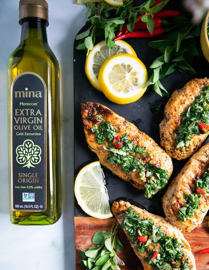 A black board with chicken chimuchurri surrounded by lemon slices, fresh oregano and a bottle of olive oil