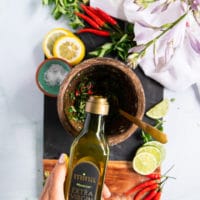 A hand pouring olive oil over the herbs and garlic to bind the chimichurri sauce