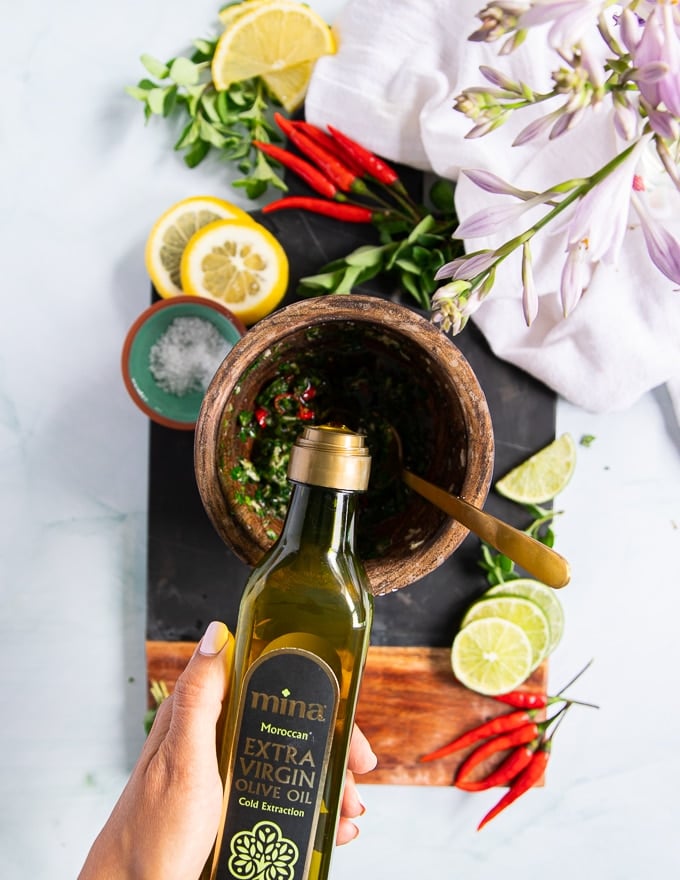 A hand pouring olive oil over the herbs and garlic to bind the chimichurri sauce