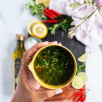 A hand holding a bowl of chimichurri recipe showing the chimichurri sauce in olive oil