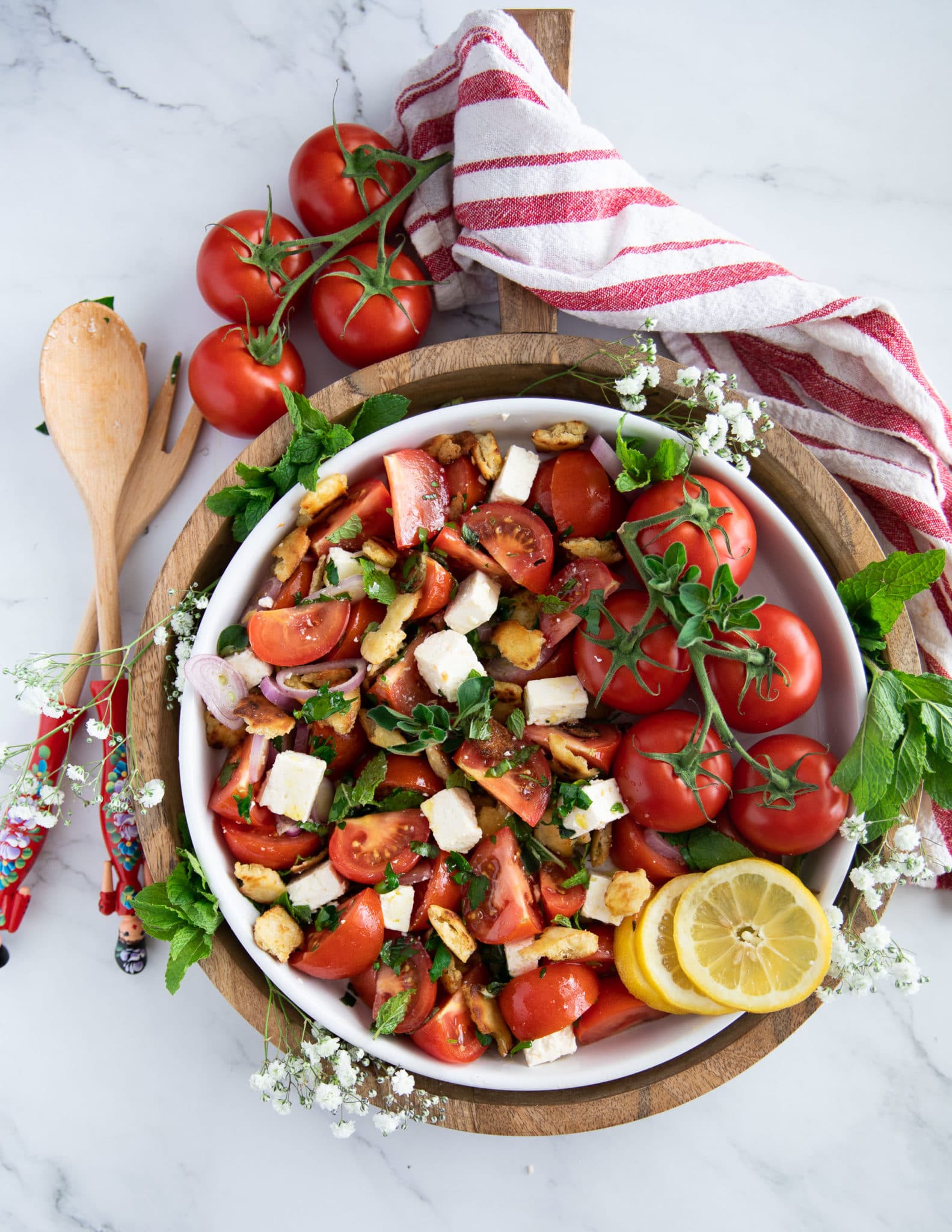 final plate of a tomato salad recipe surrounded by fresh tomatoes, herbs, lemon slices and a tea towel and two salad serving spoons