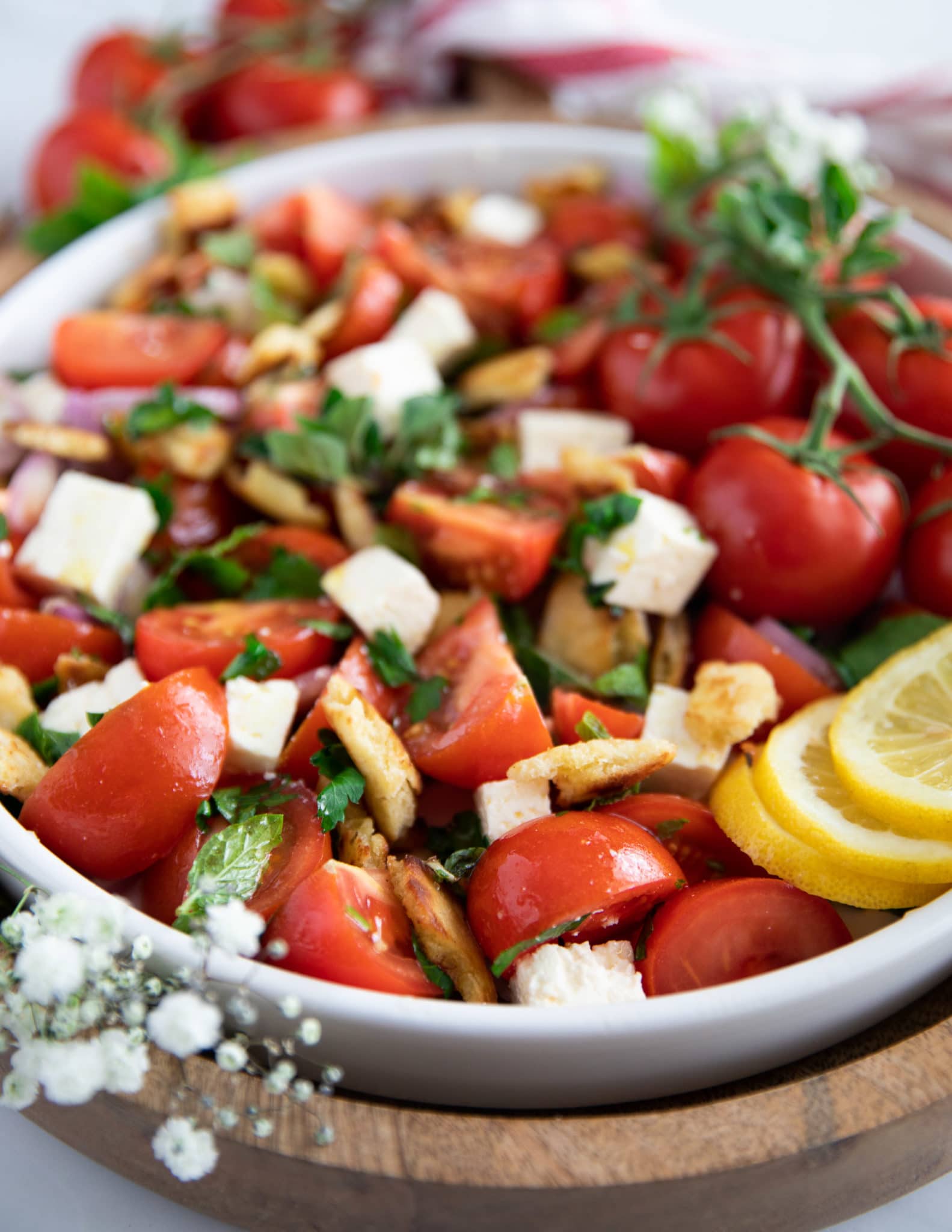 A plate of tomato salad showing the fresh herbs, feta and bread surrounded by lemon slices on a wooden board