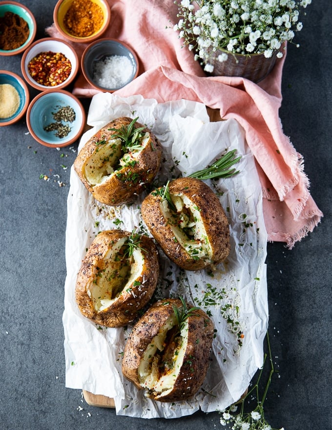 A board with four air fryer baked potatoes, cut in half and seasoned with spice and herbs, plus extra spice on the side and fresh rosemary sprigs