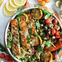 Grilled salmon on a plate with some grilled veggies, chickpea salad, surrounded by lemon slices