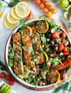 Grilled salmon on a plate with some grilled veggies, chickpea salad, surrounded by lemon slices