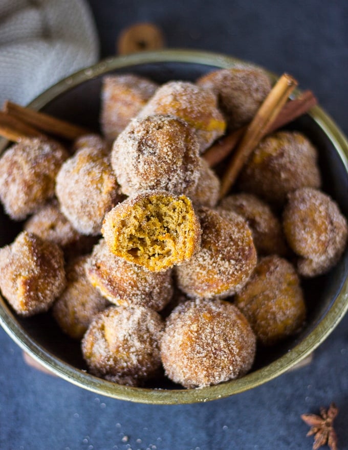 A bowl of pumpkin donuts coated in cinnamon sugar and one piece of donut is bitten to show the texture inside so moist and soft