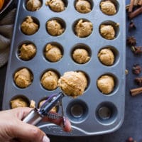 a hand holding an ice cream scoop showing how to scoop the batter into the prepared mini muffin tin for baking the donut holes