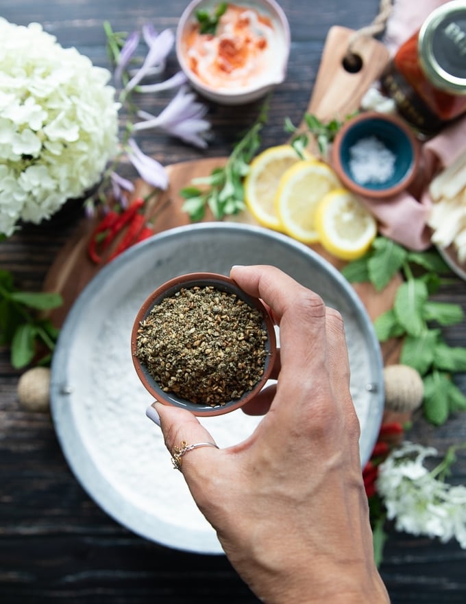 a hand holding a small bowl of zaatar spice and adding it to a bowl of flour to season the flour