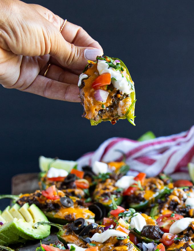 a hand holding an eaten up jalapeno popper over a plate of baked jalapeno poppers