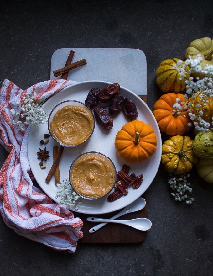 pumpkin chia pudding in individual serving bowls on a plate with walnuts, dates, a small pumpkin and cinnamon sticks. 