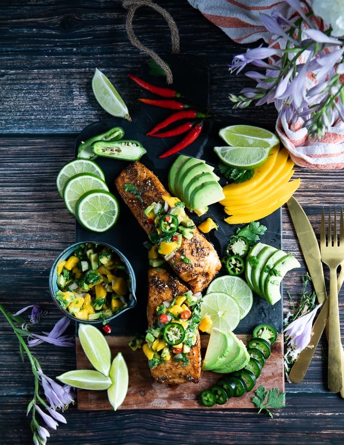 two pieces of air fryer salmon on a board surrounded by lime wedges, avocado slices, mango, avocado salsa, 