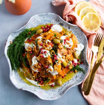 A plate of roasted honeynut squash served over yogurt sauce and topped with more yogurt sauce, harissa sauce, fresh dill, olives, pomegranates, toasted pine nuts and olive oil