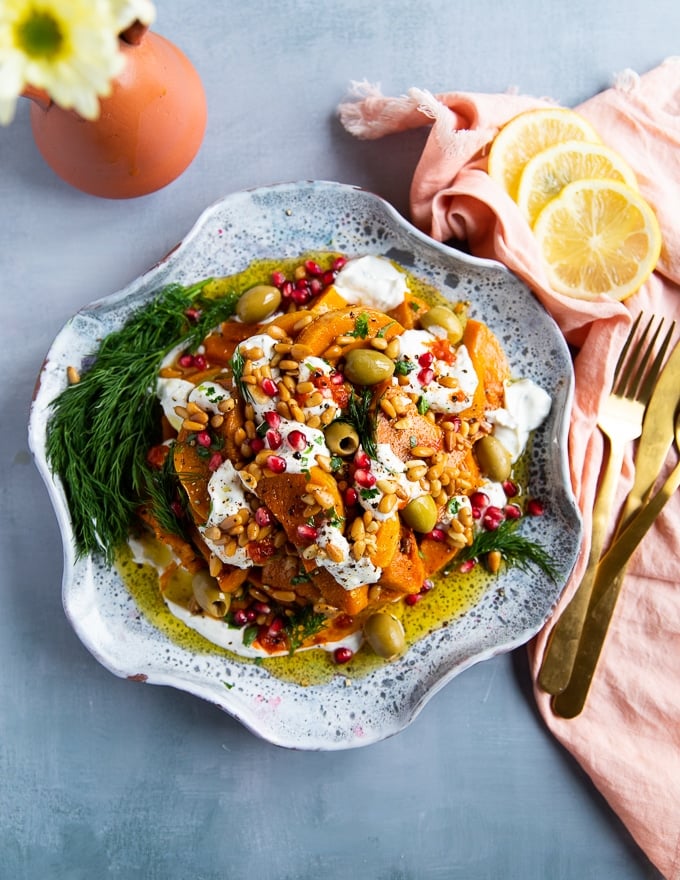 A plate of roasted honeynut squash served over yogurt sauce and topped with more yogurt sauce, harissa sauce, fresh dill, olives, pomegranates, toasted pine nuts and olive oil
