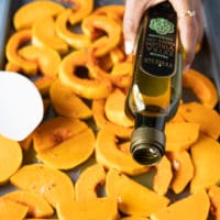 squash on a baking trat seasoned and a hand drizzling olive oil over the squash before roasting
