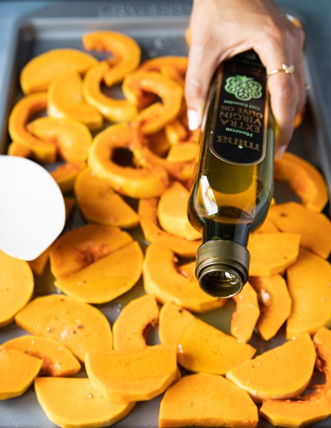 squash on a baking trat seasoned and a hand drizzling olive oil over the squash before roasting