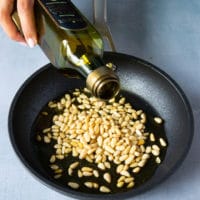 Raw Pine nuts in a skillet and a hand pouring over some olive oil over the nuts