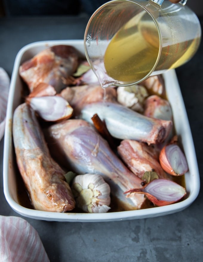 a hand pouring stock over the lamb shanks with whole spices and onions in the baking dish to roast