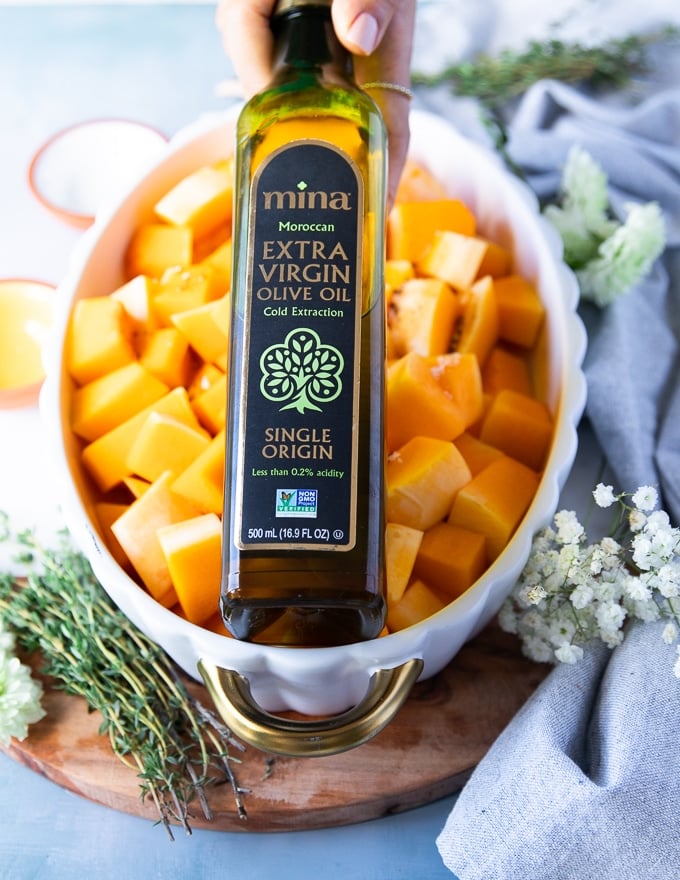 a hand holding olive oil drizzling it over the but up butternut squash in a casserole dish