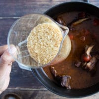 A hand holding a cup of rice adding it to the pan of the cooked lamb shoulder chops along with stock