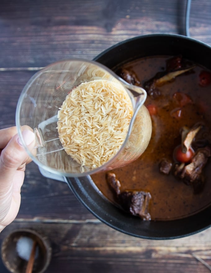 A hand holding a cup of rice adding it to the pan of the cooked lamb shoulder chops along with stock