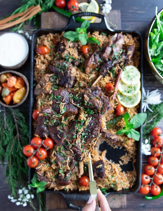 A hand holding off a piece of cooked lamb shoulder chops over the plate of rice and lamb