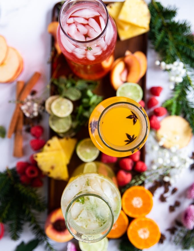 three bottles of mocktails made and ready to scoop surrounded by garnishes of fresh fruit, herbs, and whole spice