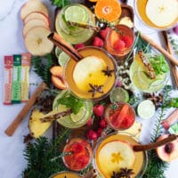 a large tray with a collection of al the mocktail recipes and different garnishes such as fresh raspberries, apples, pineapple slices, fresh mint, lime, fresh thyme and whole spices