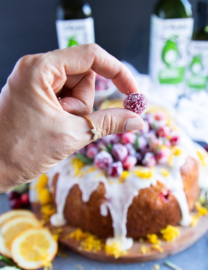 A hand holding a sugared cranberry ready to add them over the orange cake