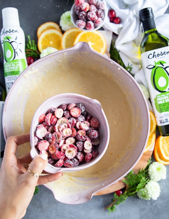 a hand holding the bowl of flour tossed cranberries over the batter bowl ready ot add it in for the orange cake 