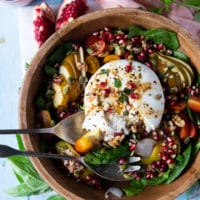a large bowl of salad and a burrata cheese over the top with pomegranate arils for garnish and two serving spoons