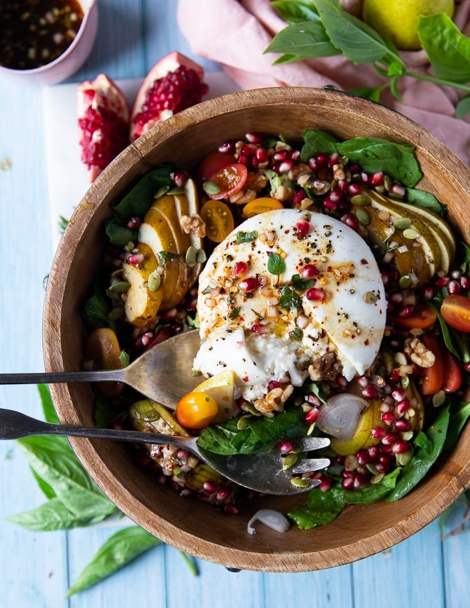 a large bowl of salad and a burrata cheese over the top with pomegranate arils for garnish and two serving spoons