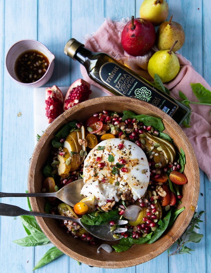 a large bowl of salad and a burrata cheese over the top with pomegranate arils for garnish and two serving spoons surrounded by fresh pears, more dressing and a bottle of olive oil