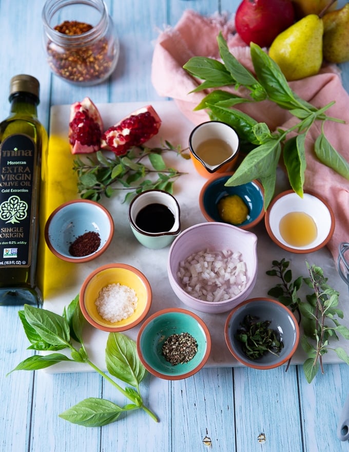 salad dressing ingredients including olive oil, a bowl of shallots, pomegranate molasses, vinegar, seasoning, honey, herbs, mustard