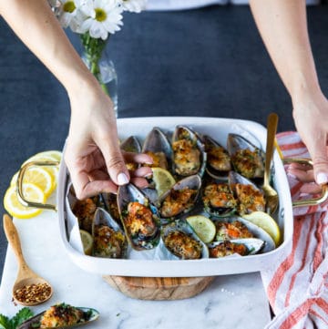 A hand holding a green mussel over a tray of cooked new zealand mussels to show the size and texture