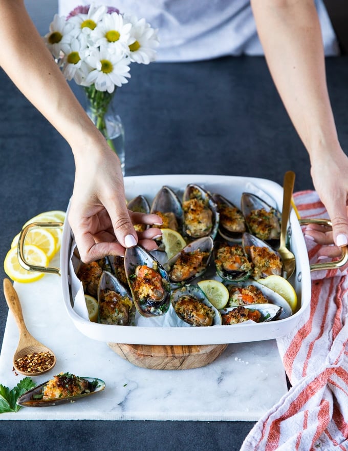 A hand holding a green mussel over a tray of cooked new zealand mussels to show the size and texture 
