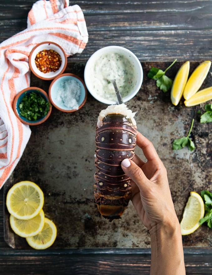 A hand holding a fresh raw lobster tail surrpunded by garlic sauce, fresh herb,s lemon juice, seasoning and melted butter