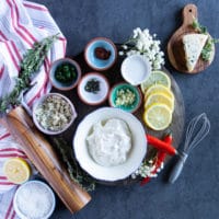 All blue cheese ingredients on a wooden board surrounded by a tea towel. A bowl of sour cream, a wedge of blue cheese, lemon, garlic, fresh herbs, salt and pepper and milk