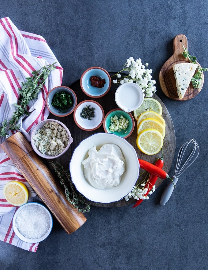 All blue cheese ingredients on a wooden board surrounded by a tea towel. A bowl of sour cream, a wedge of blue cheese, lemon, garlic, fresh herbs, salt and pepper and milk 