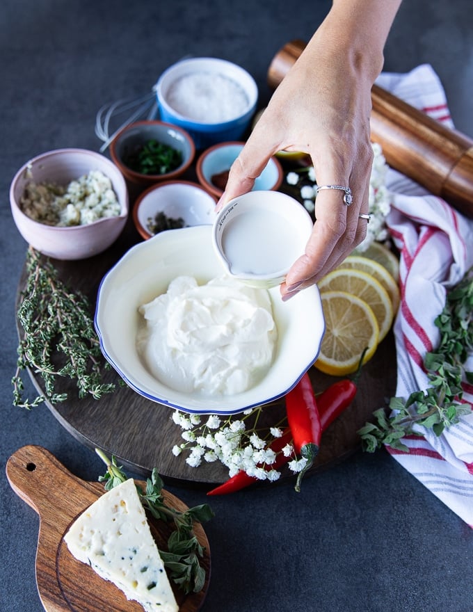 A hand holding a small cup of milk pouring it into a bowl of blue cheese dressing to adjust consistency of the dressing