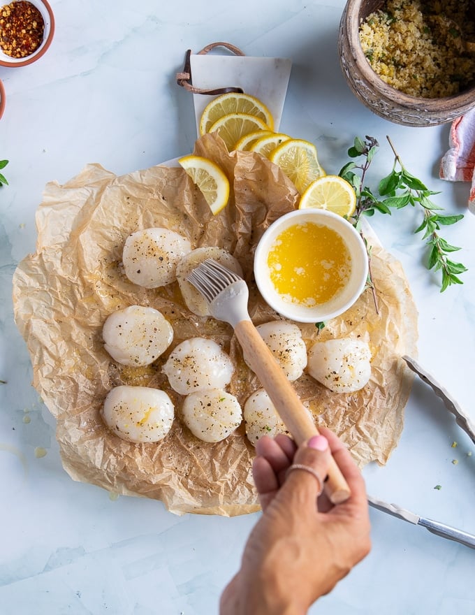 A hand brushing the scallops with melted butter preparing them to bake