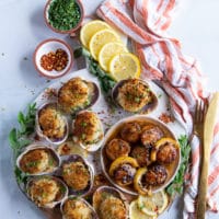 A board with two scallops recipes, a plate of seared scallops and a plate of baked scallops surrounded by lemon slices and a tea towel showing the different ways of how to cook scallops